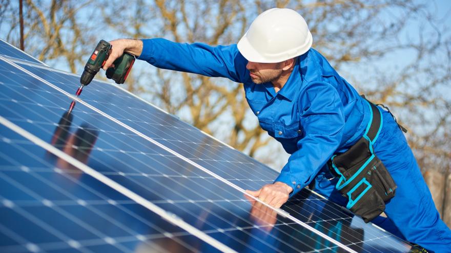 Arbeiter in blauem Anzug und Schutzhelm bei der Installation einer Photovoltaikanlage mit einem Schraubenzieher. 